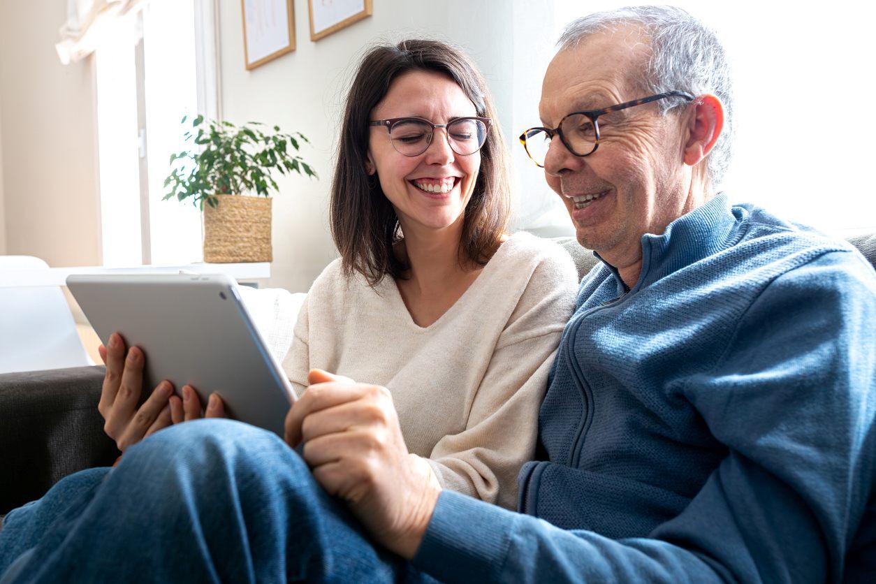 A smiling senior father and adult daughter look at an iPad.