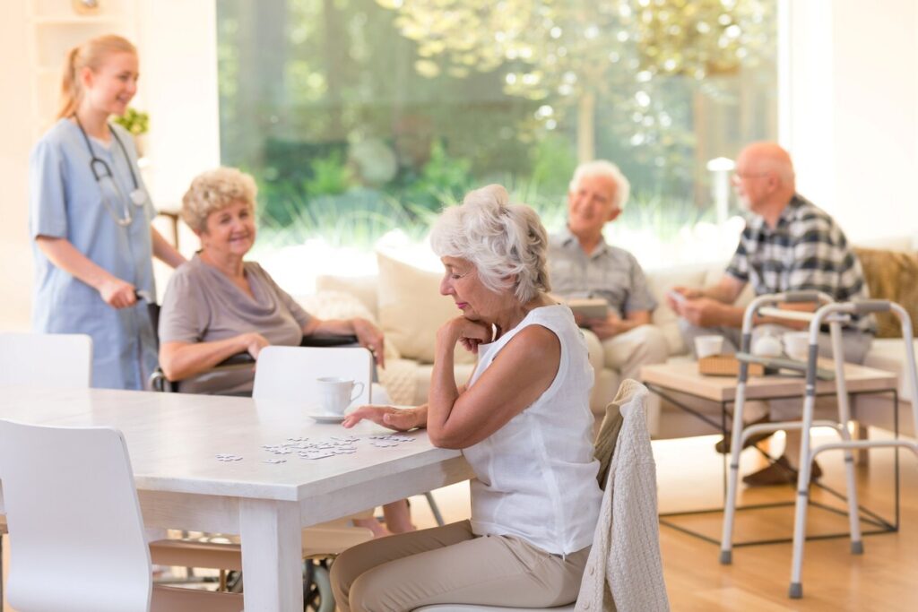 A senior woman sits at a table doing a puzzle with seniors and a nurse behind her.