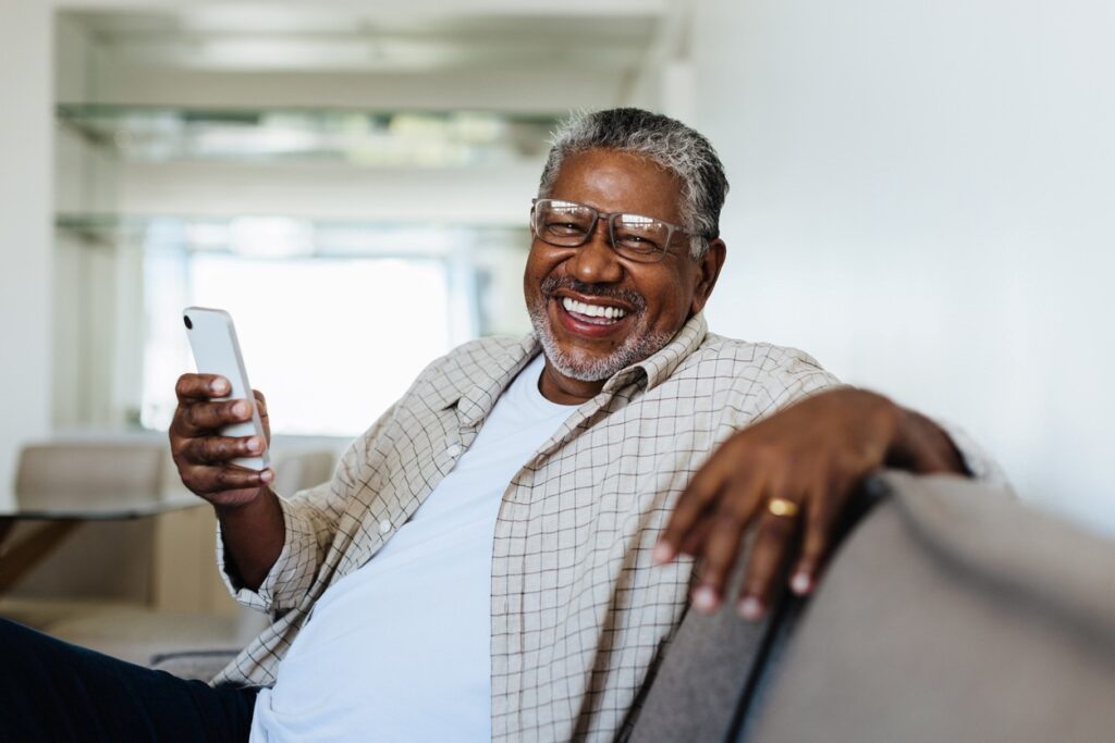 senior man smiles as he sits on a couch, looking at his smartphone.