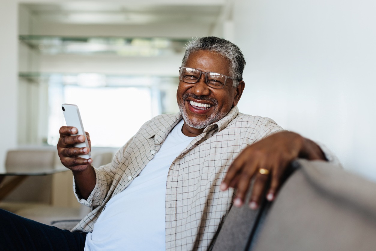 senior man smiles as he sits on a couch, looking at his smartphone.