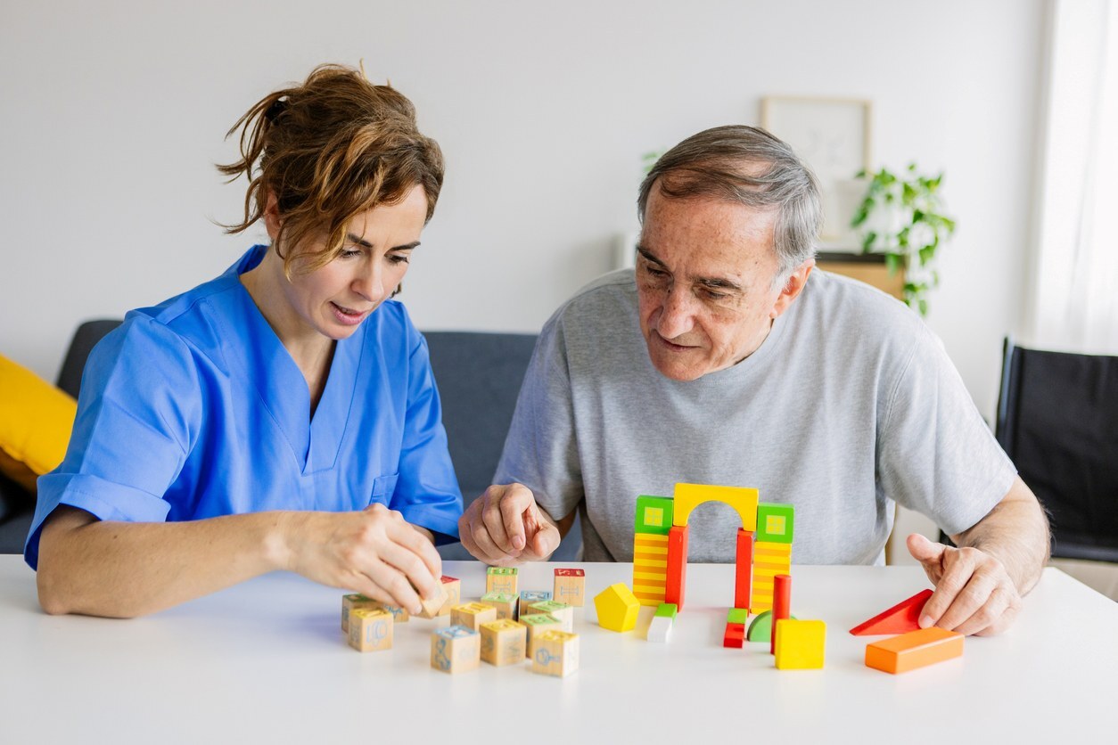 A caregiver and a senior play a wooden block puzzle on a table in a white room with a plant on the wall.