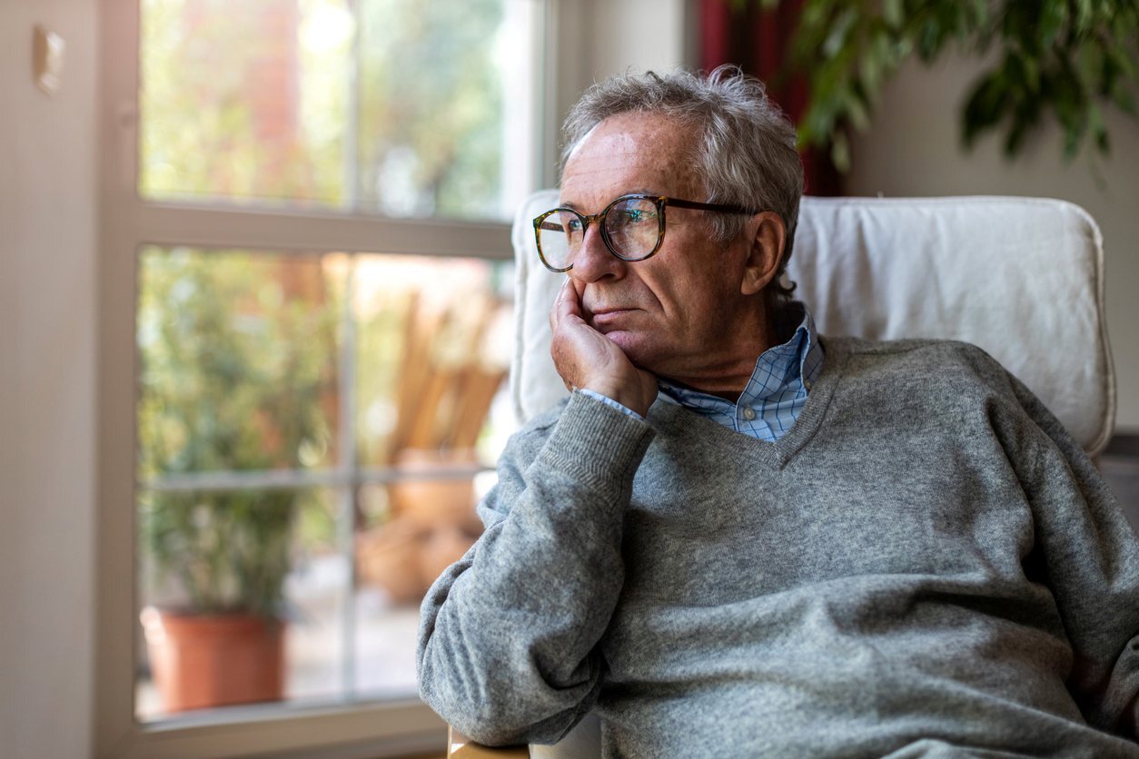 An older man with his hand under his chin stares out of the window at home.
